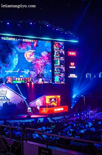 A large esports arena featuring a massive screen displaying a video game, surrounded by sponsor logos and team names. The auditorium is filled with spectators watching the gaming event. Neon blue and purple lights illuminate the stage, creating a vibrant atmosphere.